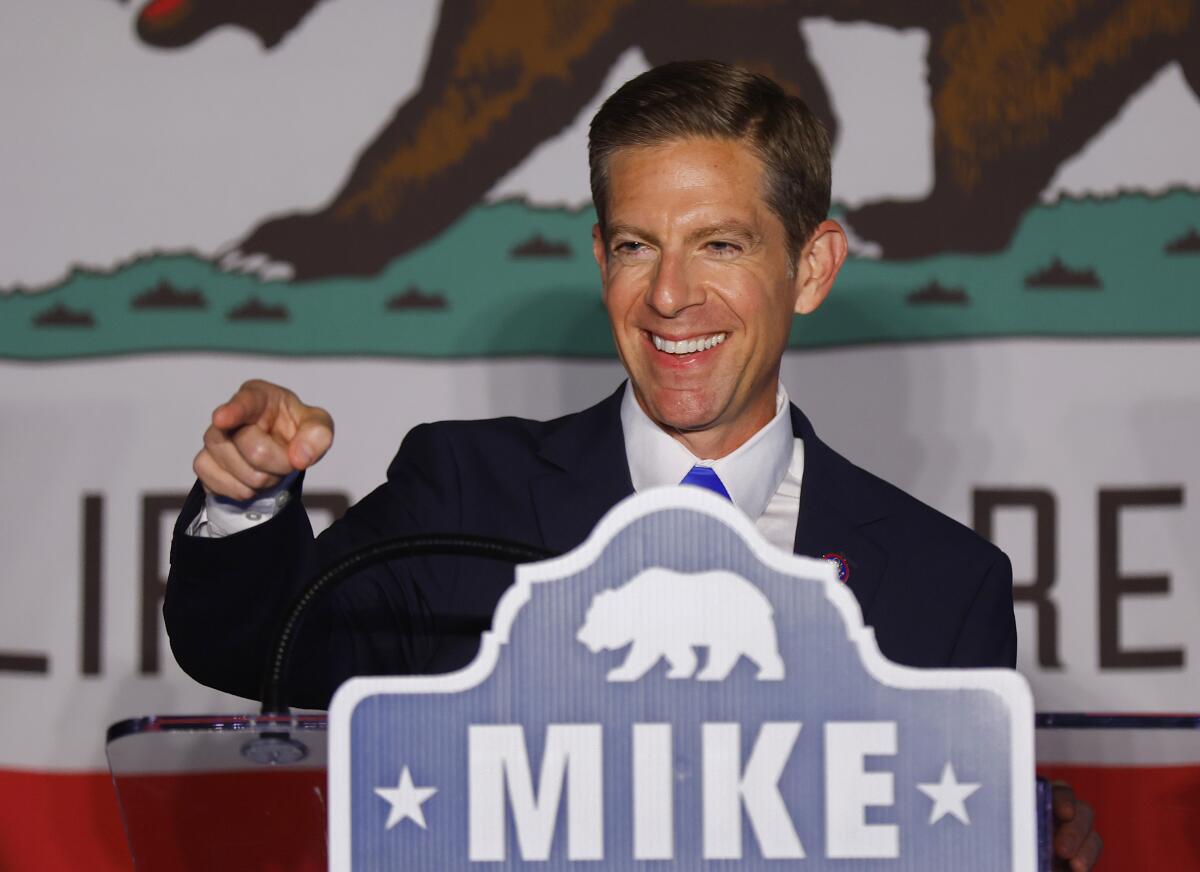 A smiling man in a suit, standing at a podium labeled "Mike" in front of a California flag, points out of the frame.