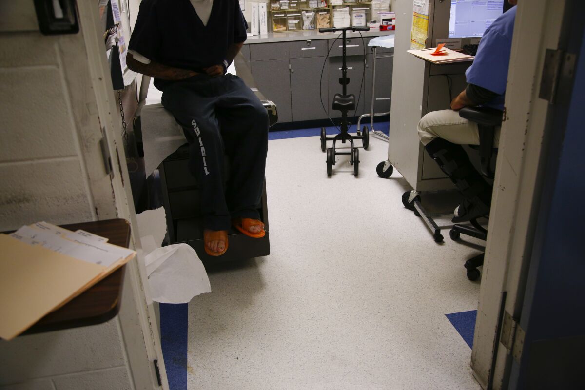 An incarcerated man in a jumpsuit sits in a jail medical unit for a check-up. 