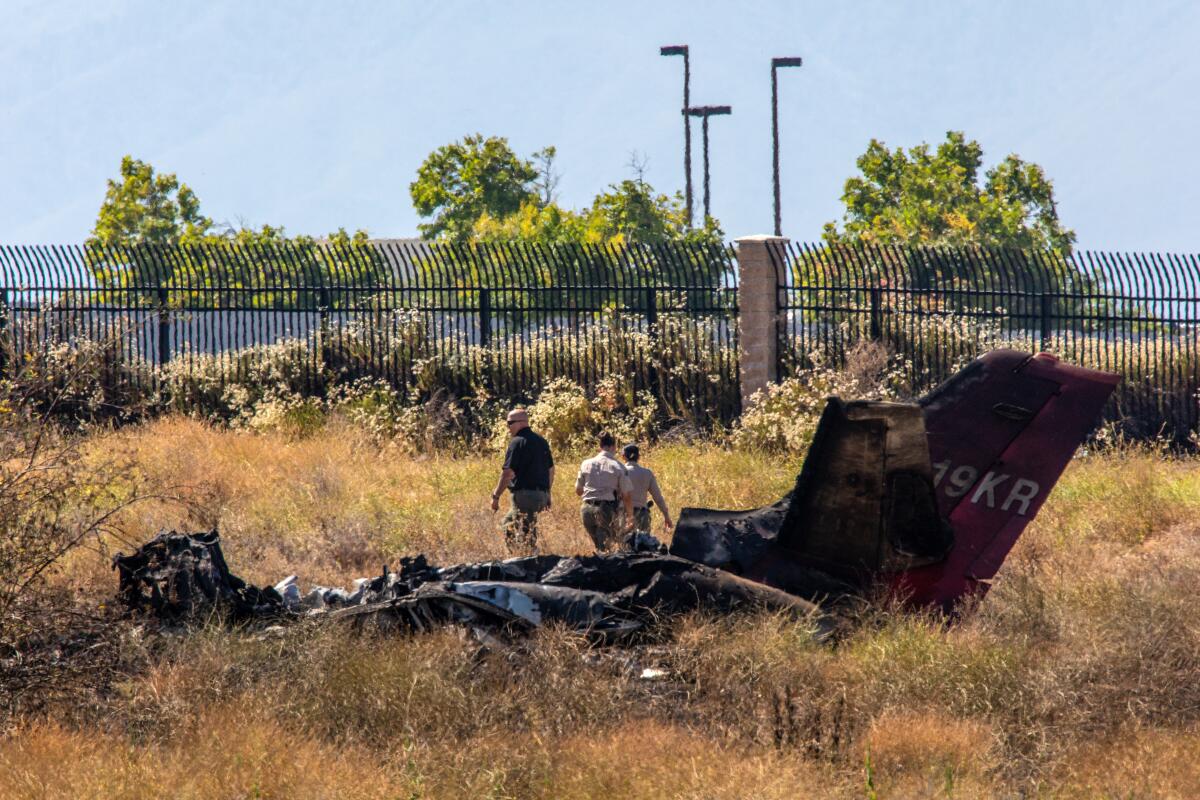 Part of a fire-damaged plane rests in a field with three people behind it.