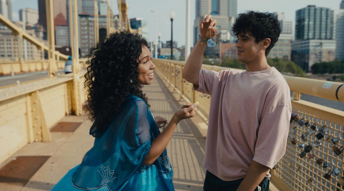 A young man shows his girlfriend a small blue heart on a chain as they stand on a bridge.