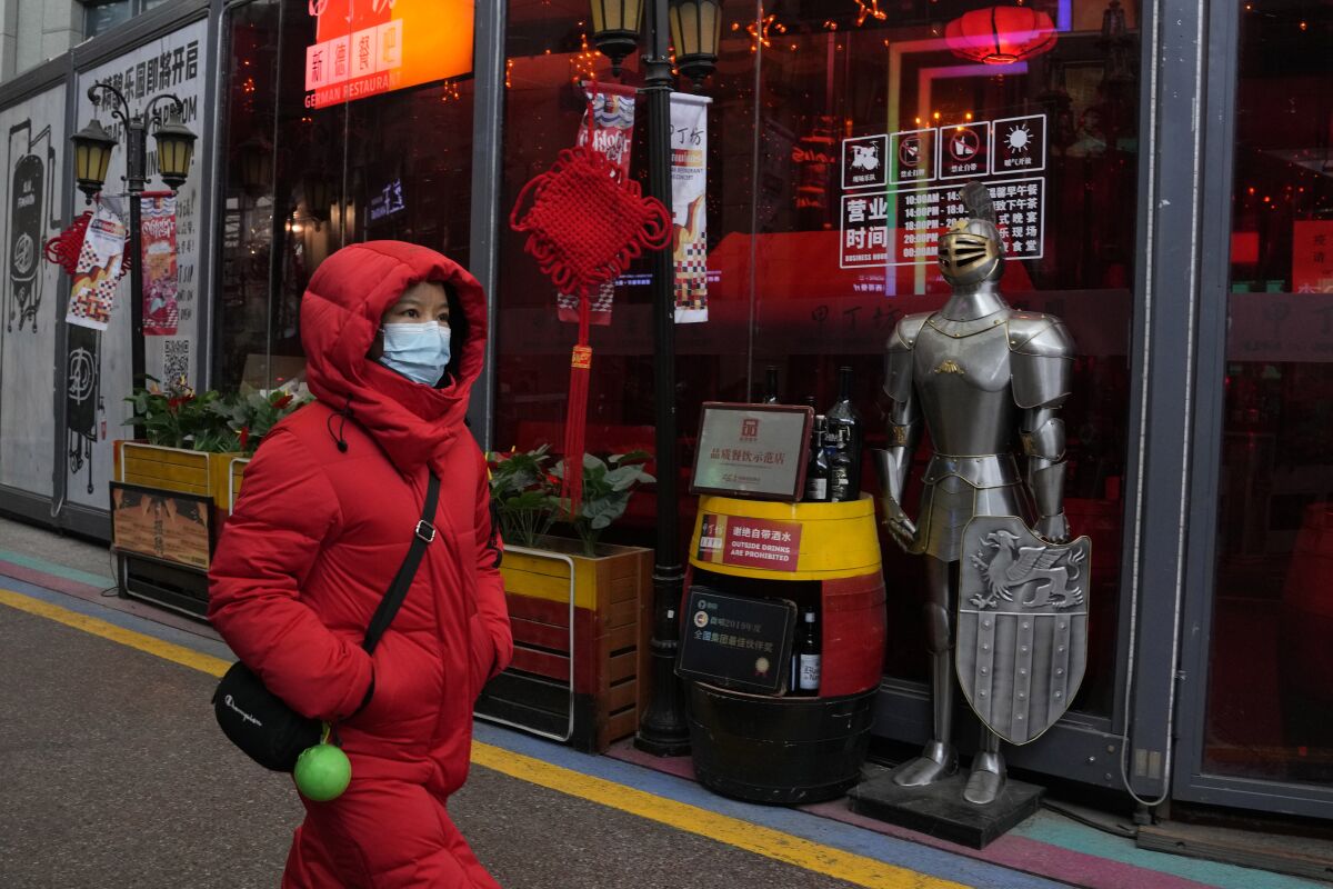 A woman wearing a mask to protect from the coronavirus walks past a coat of armor displayed outside a restaurant in Beijing.