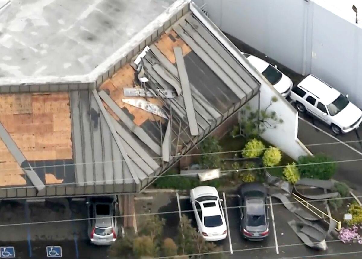 An aerial frame of an industrial building with damaged roof and debris strewn near parked cars.