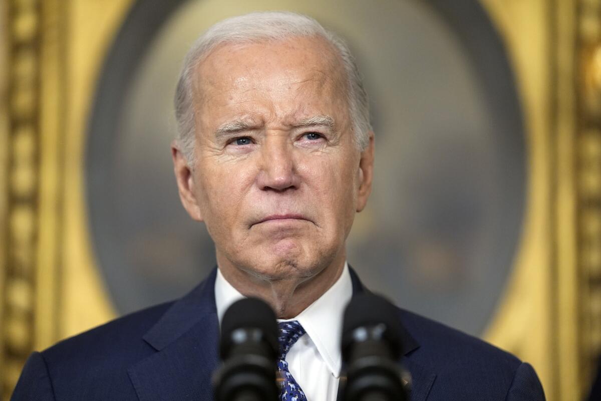 A closeup of President Biden in front of two microphones against a gilded background