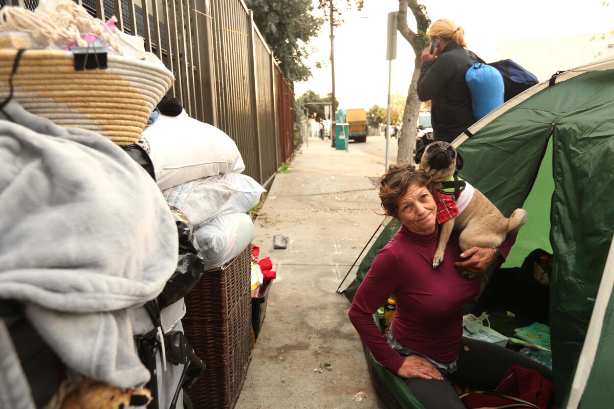 A woman holding a small dog sits in the entrance to a tent on a sidewalk