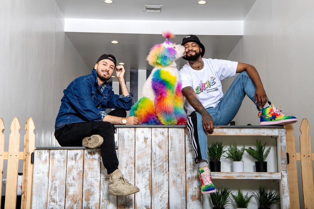Two men sit on a countertop with their tye-dyed dog in the middle, who is kissing the man to the right