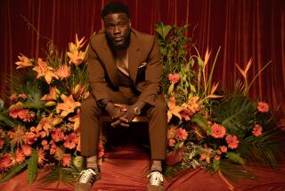 Kevin Hart sits on a chair surrounded by orange flowers and leans forward onto his knees 