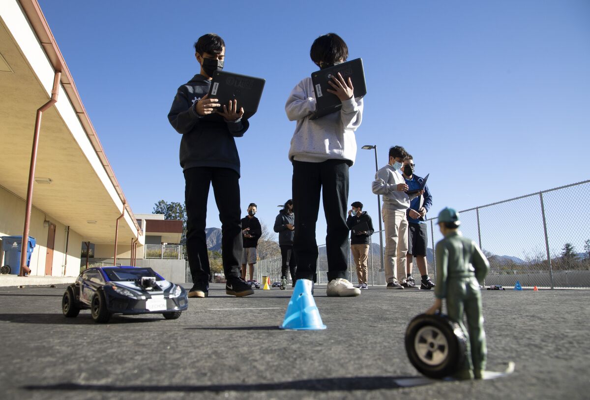 Two students wear face masks and hold clipboards as they tower above a toy car and figure of a mechanic on a school blacktop.