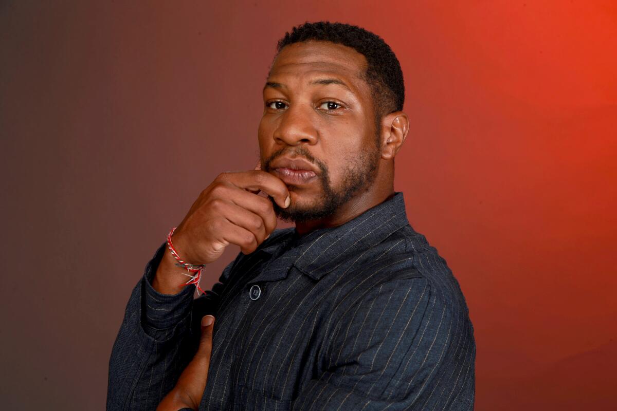 Jonathan Majors holds his hand to his chin while posing against a plain red-orange backdrop