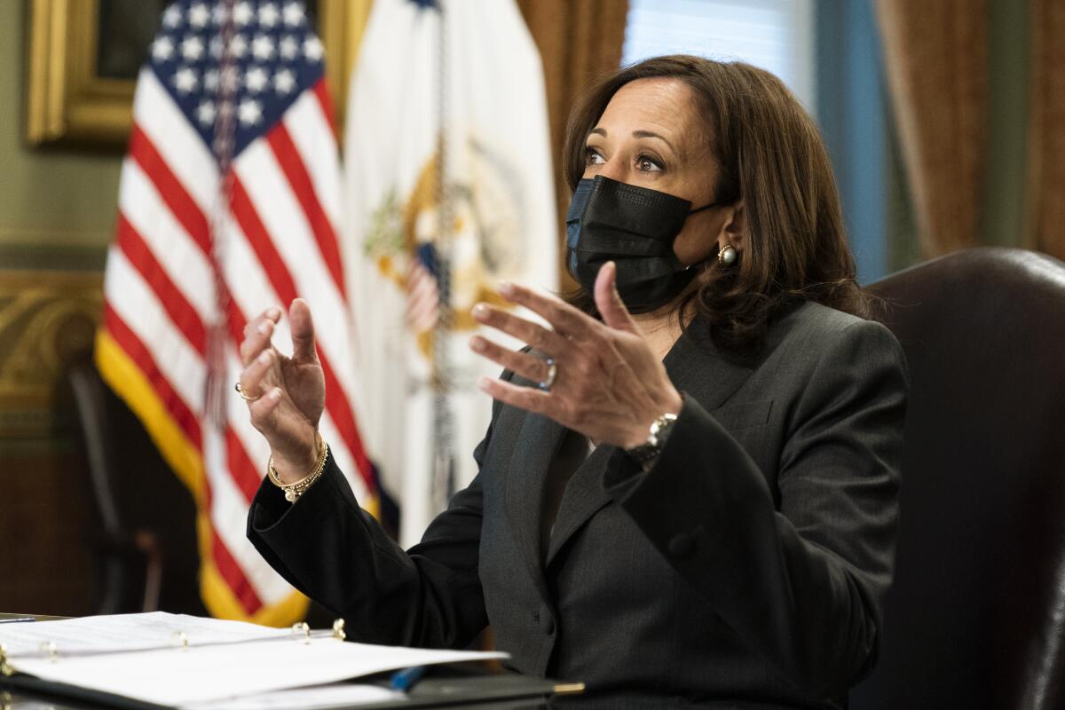 Vice President Kamala Harris wears a mask and gestures while sitting at her desk.