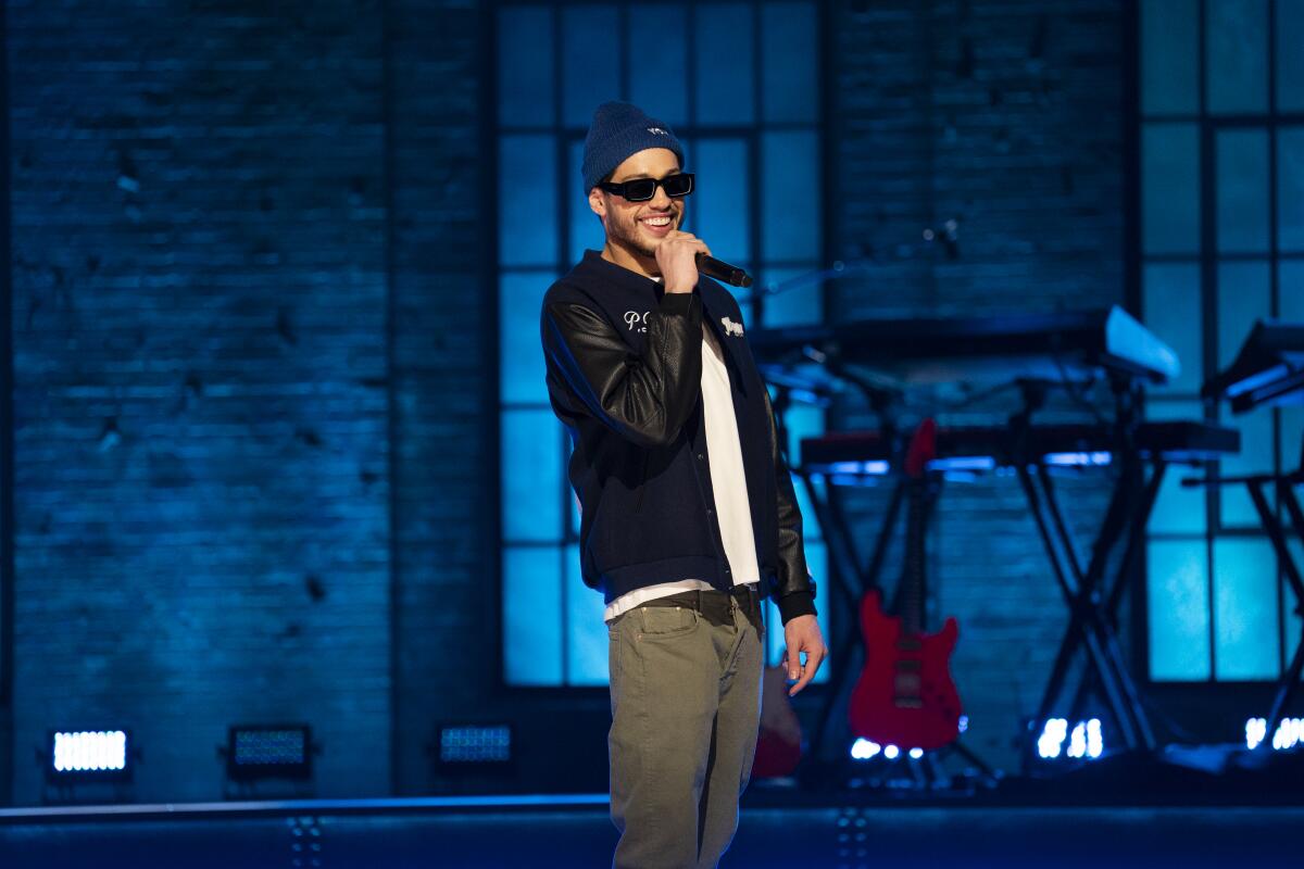 A man wearing a beanie and sunglasses and speaking into a microphone on a stage