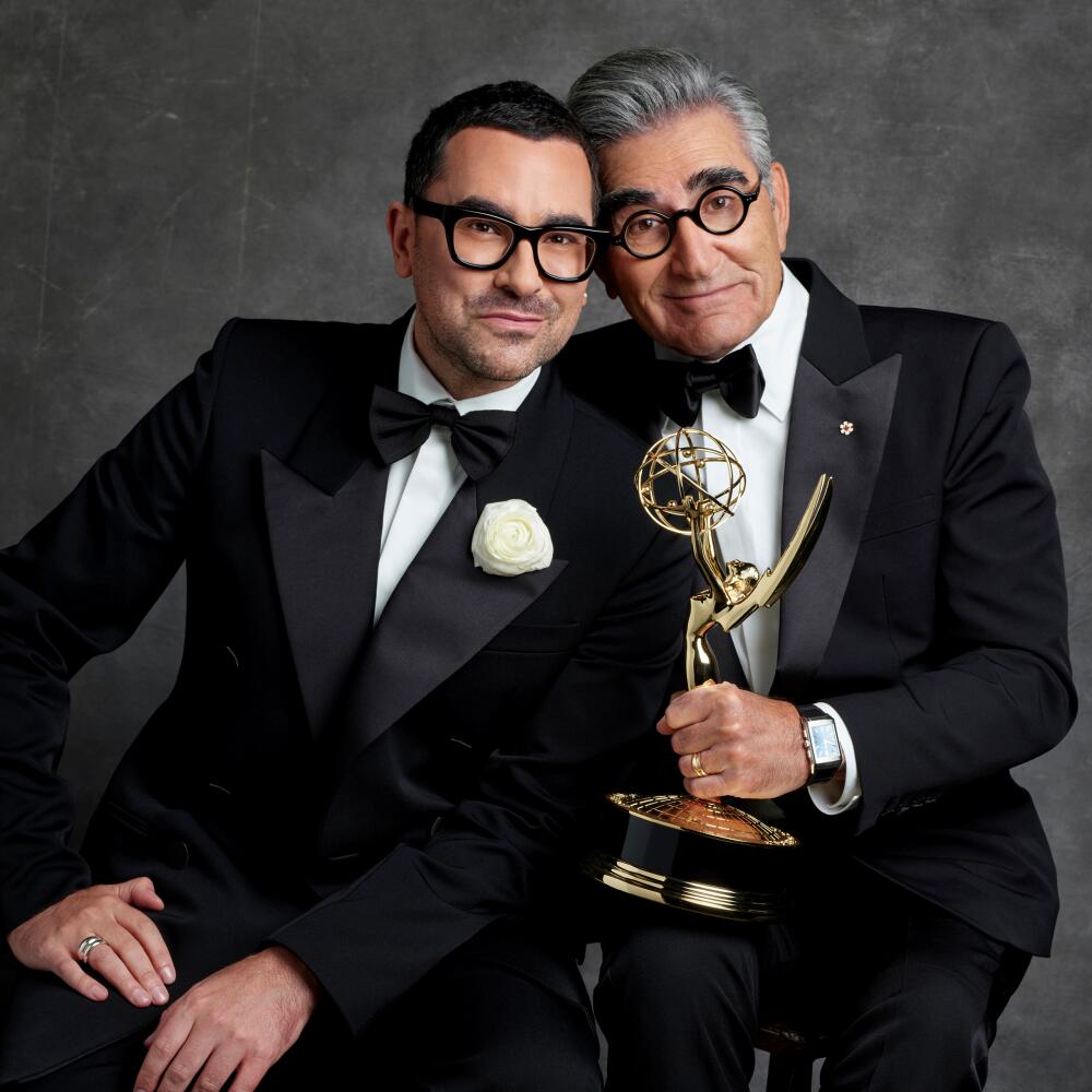 Two men in tuxedos and dark-rimmed glasses, one holding an Emmys statuette, sit and lean against each other.