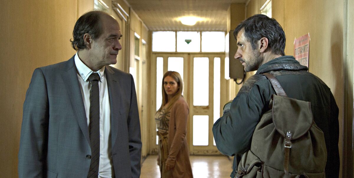 A woman watches as a man in a suit faces off with a man with a rucksack in a hallway in the 2020 drama “Father.”