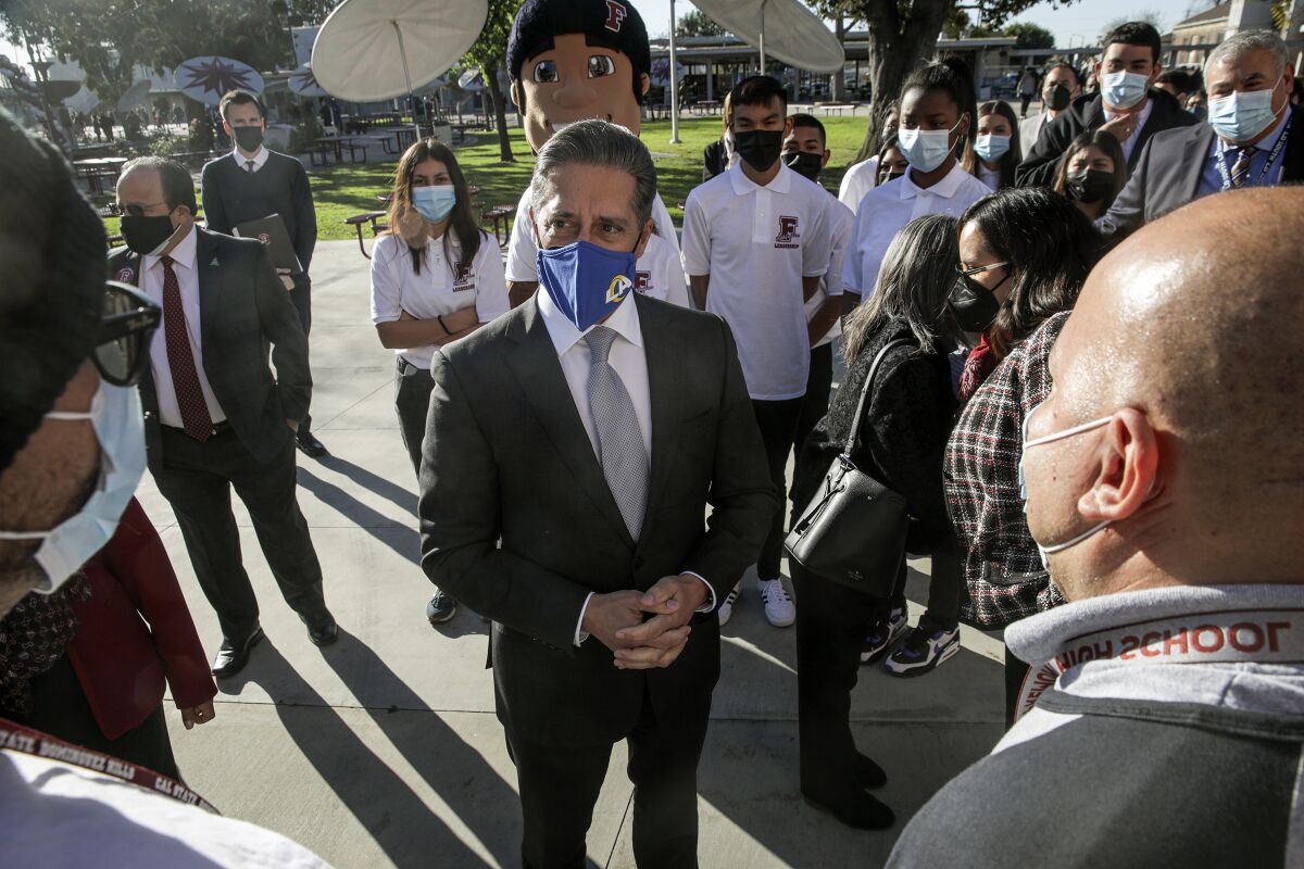 A masked man in a suit and tie stands, hands clasped, as he speaks to a group of people gathered around him