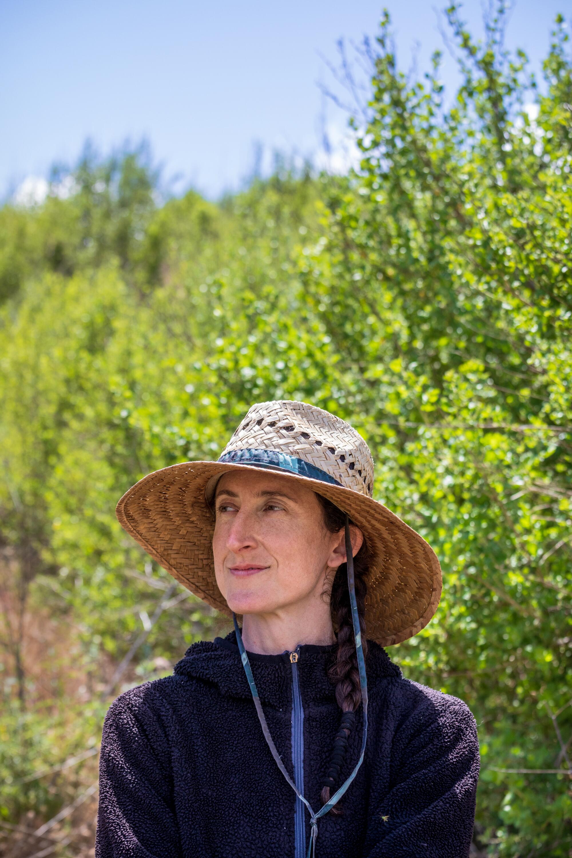 A woman wearing a broad-brimmed straw hat stands outdoors in front of a green hillside.
