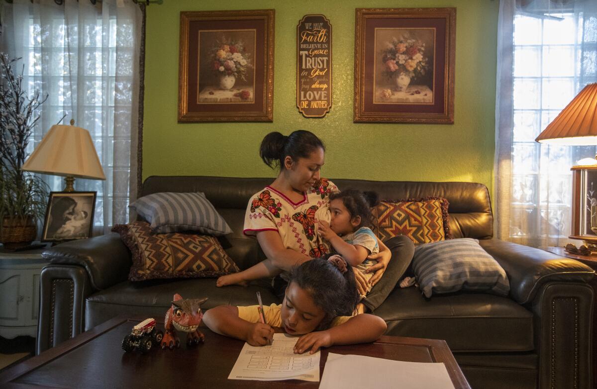 A woman nurses a child on a sofa as an older child does homework on a table in front of them.