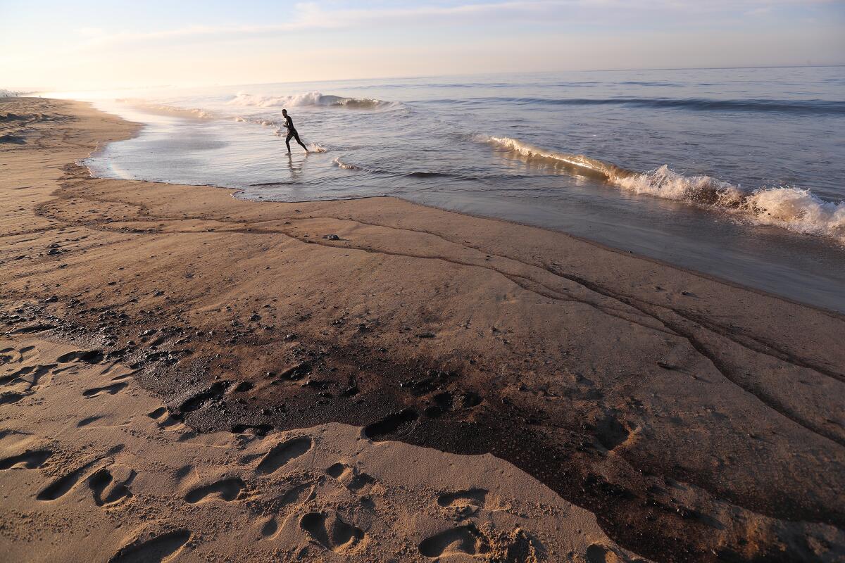 A person walks along the shore, marked by black lines of oil washing up from the ocean