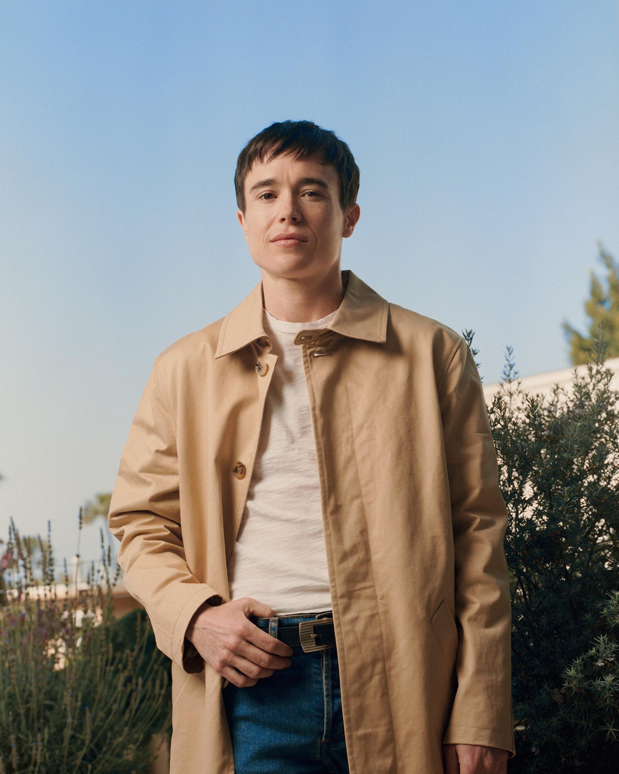 Elliot Page, wearing a tan coat, stands outdoors against a blue sky