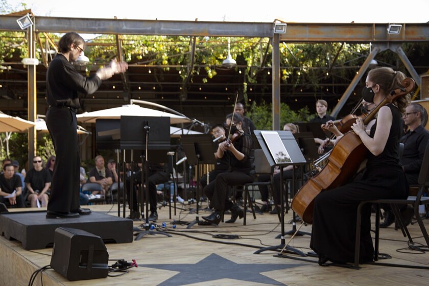 A conductor and orchestral players outdoors.