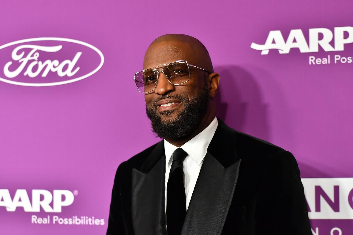 A bald man with a beard smiles while wearing sunglasses and a suit.