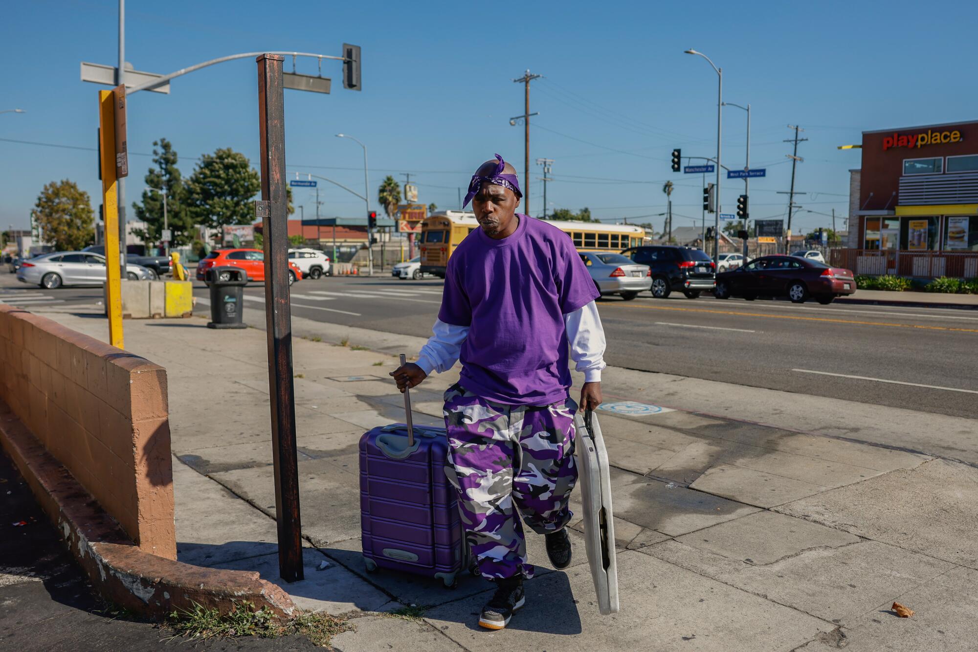 Vincent Hubbard, dress in purple, pulls along a suitcase and carries a folding table as he walks on the sidewalk. 