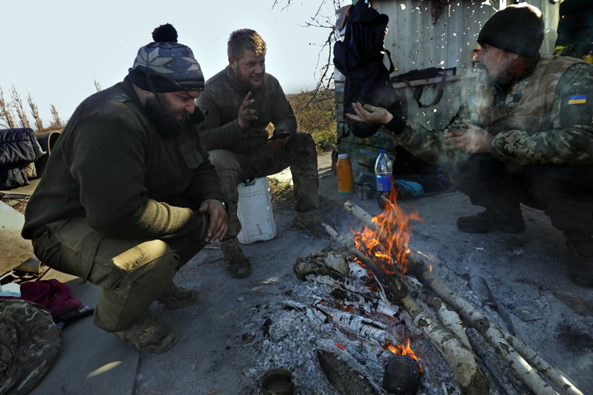 Three men in fatigues, some on their haunches, gather around a fire 