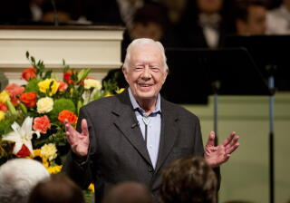 A smiling man, in a dark jacket and blue shirt, holds up his hands while standing in a church