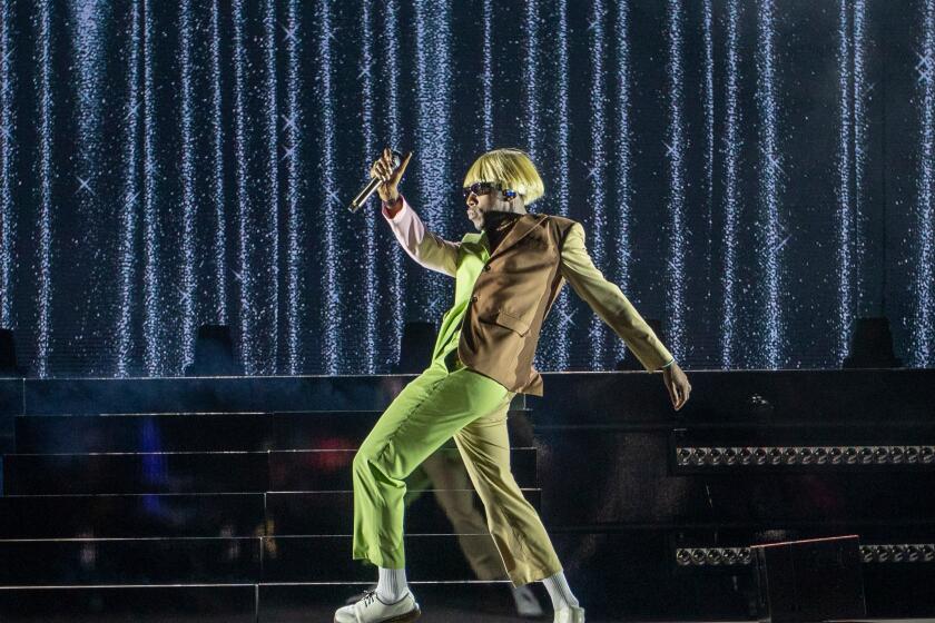 Tyler, the Creator in a platinum blonde bowl cut wig and a green an brown suit posing on a stage
