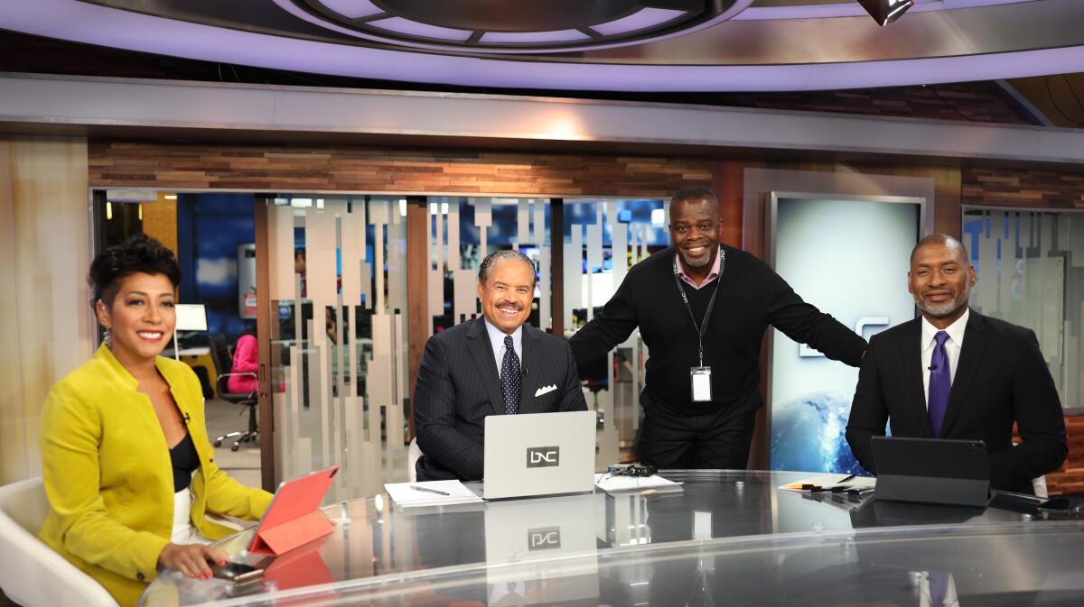 A woman and three men at a TV news desk.