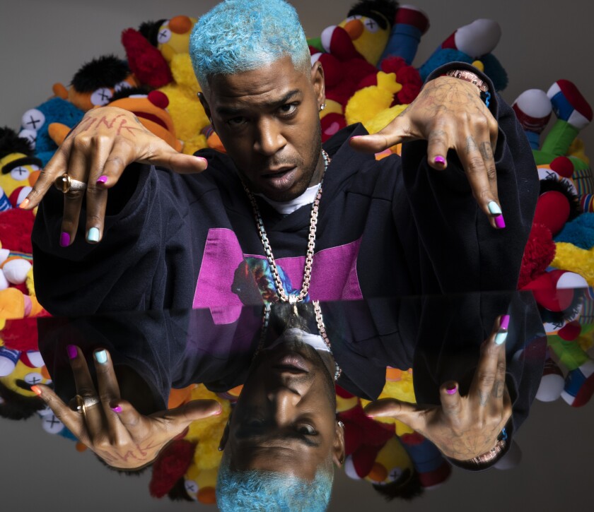 Kid Cudi, hair dyed blue and nails painted blue and fuscia, sits in front of a pile of stuffed Bert and Ernie dolls.