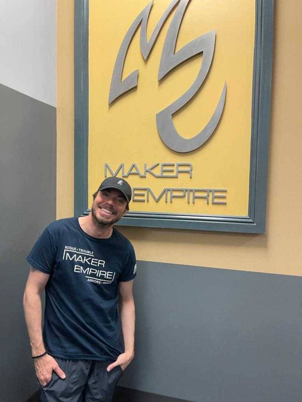 A man in a baseball cap smiles in front of a sign that reads "Maker Empire."