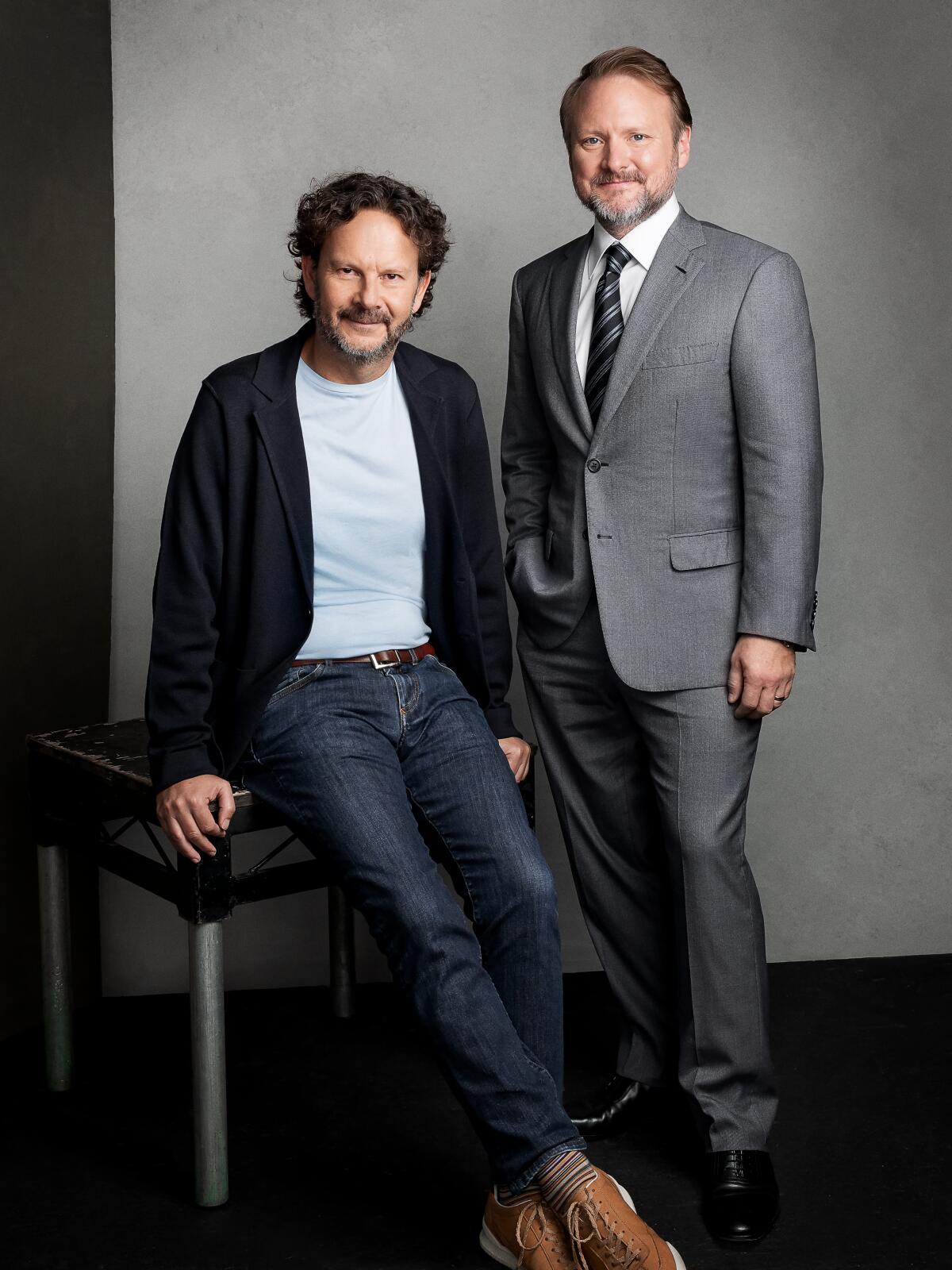 Ram Bergman sits on a stool as Rian Johnson, in a suit, stands next to him for a portrait.