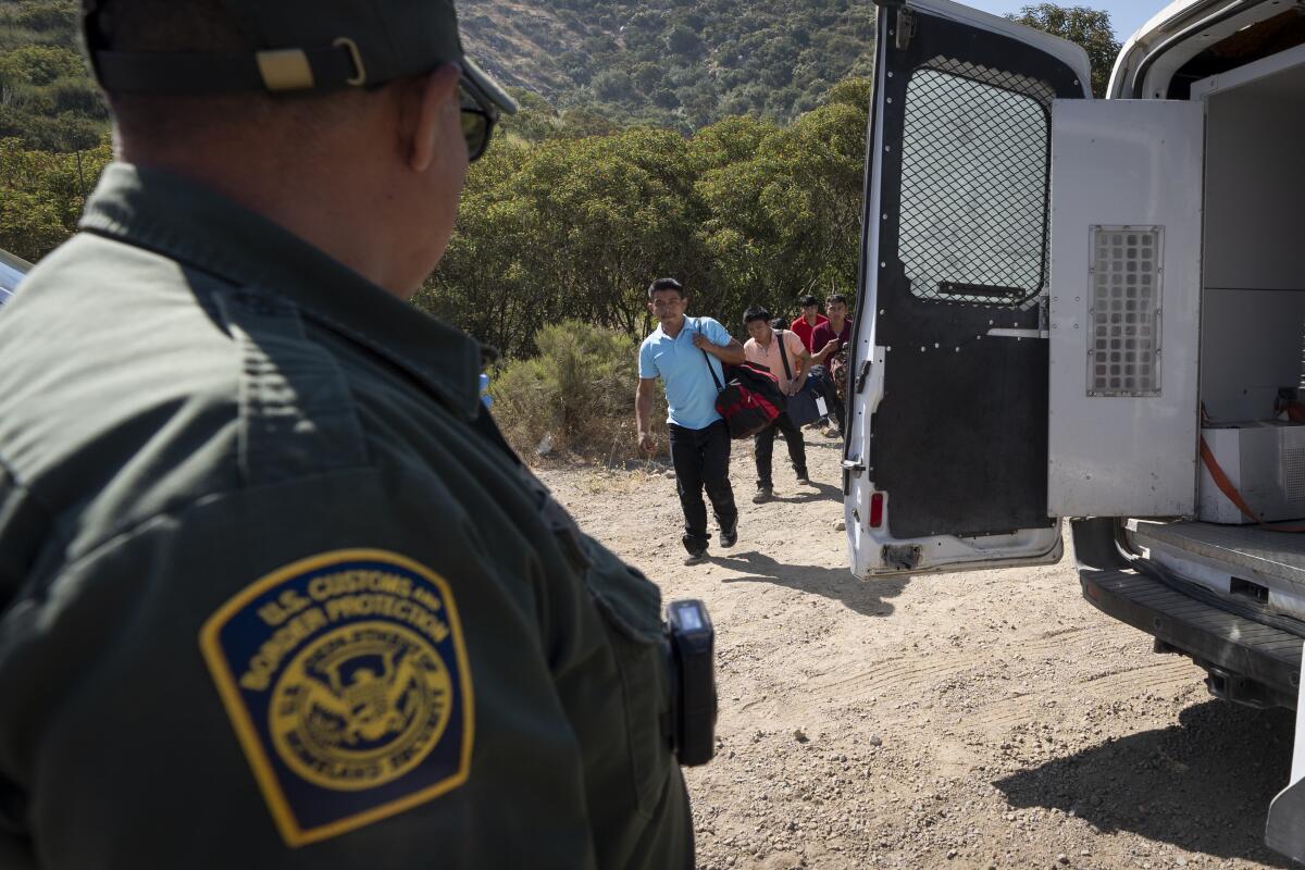 A Border Patrol agent leads a group of migrants seeking asylum towards a van to be transported and processed 