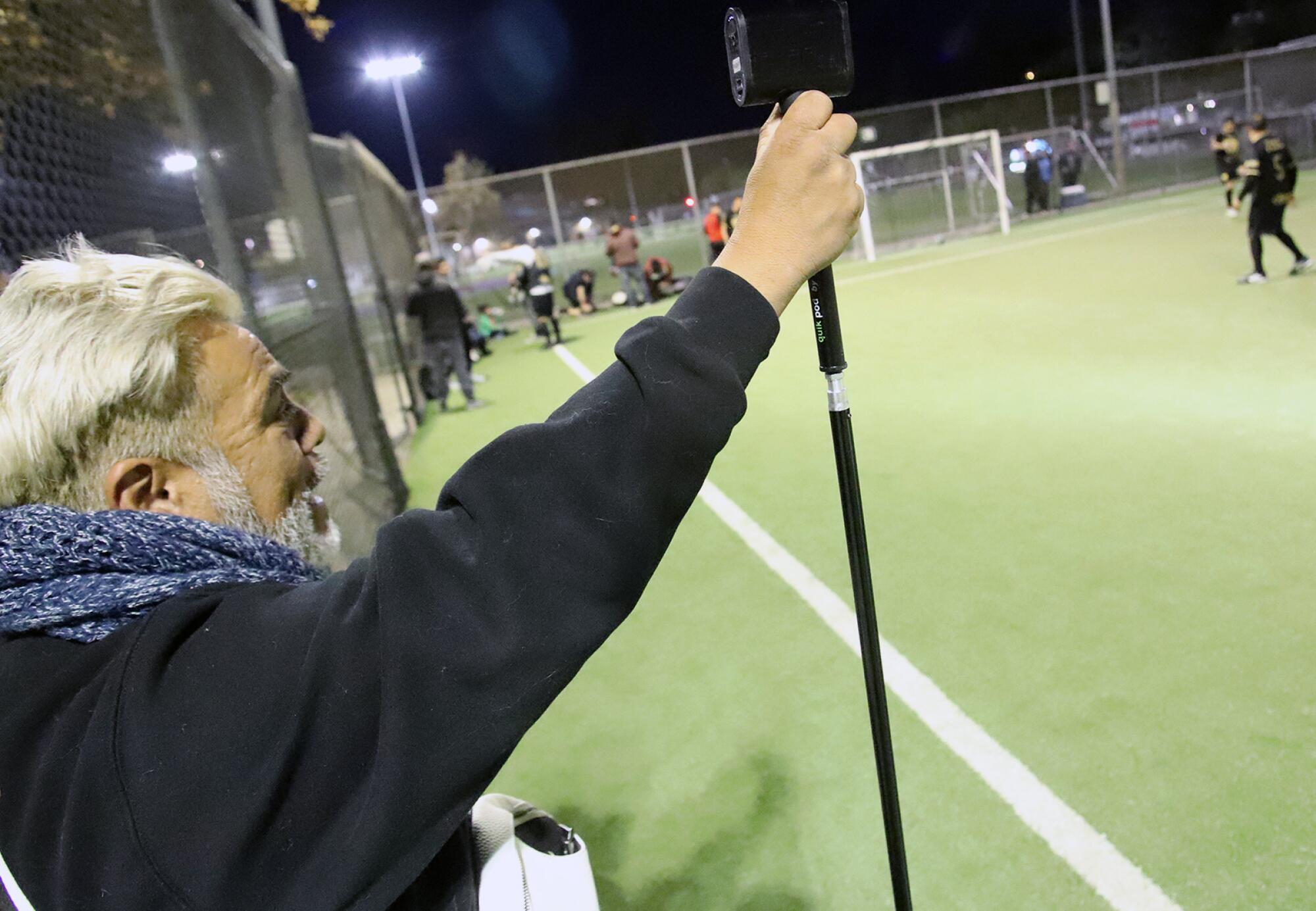 A man holds a tripod on the sideline of a soccer game.