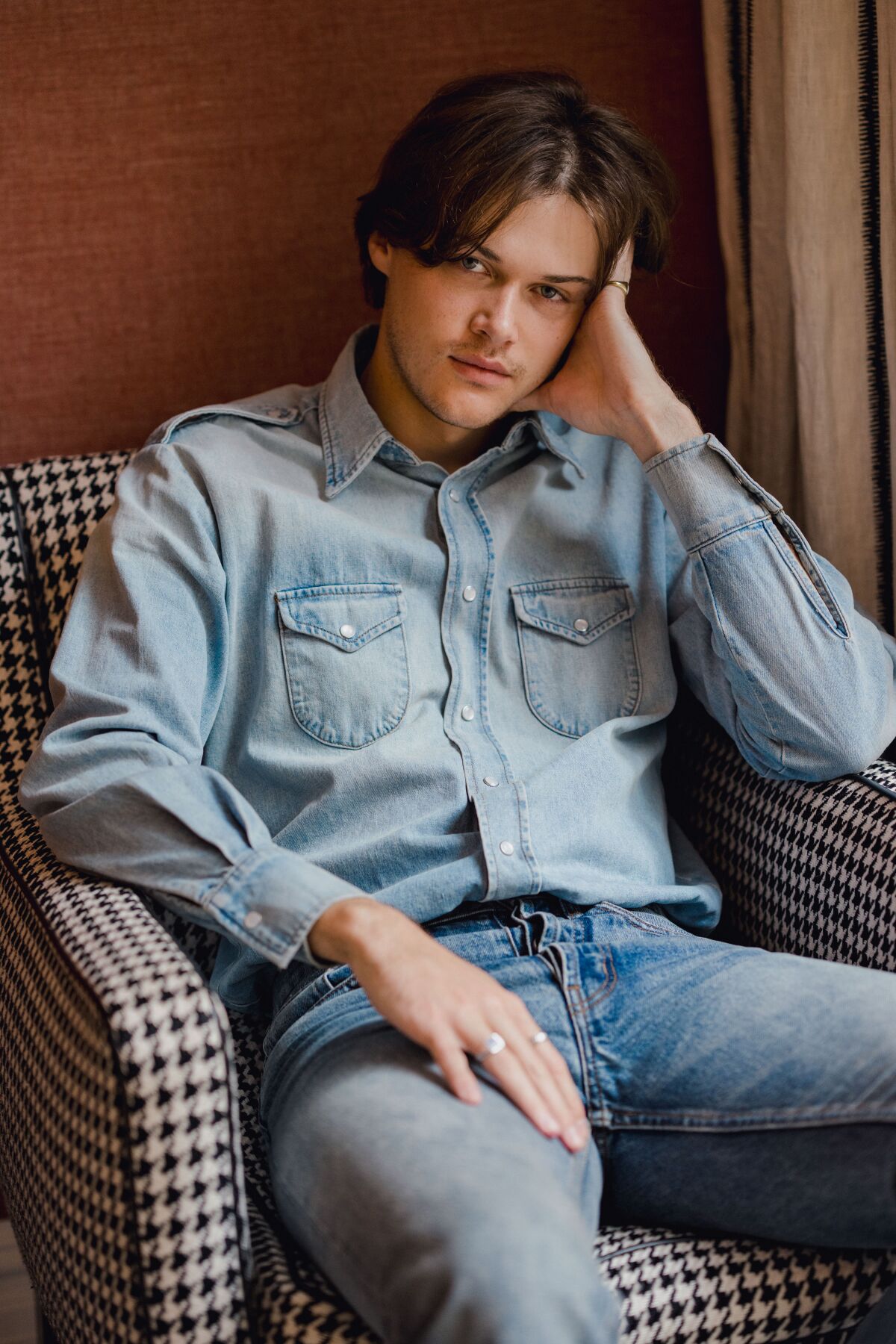 Christopher Briney in a denim shirt and jeans sits a chair.