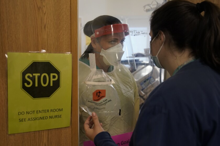 Seorang perawat yang mengenakan topeng dan wajah dan gaun pelindung menghadap orang bertopeng lain, dipisahkan oleh layar kaca sebuah ruangan