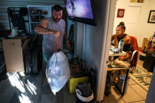 A man holds a cinched white trash bag as another person sits at a desk in another room