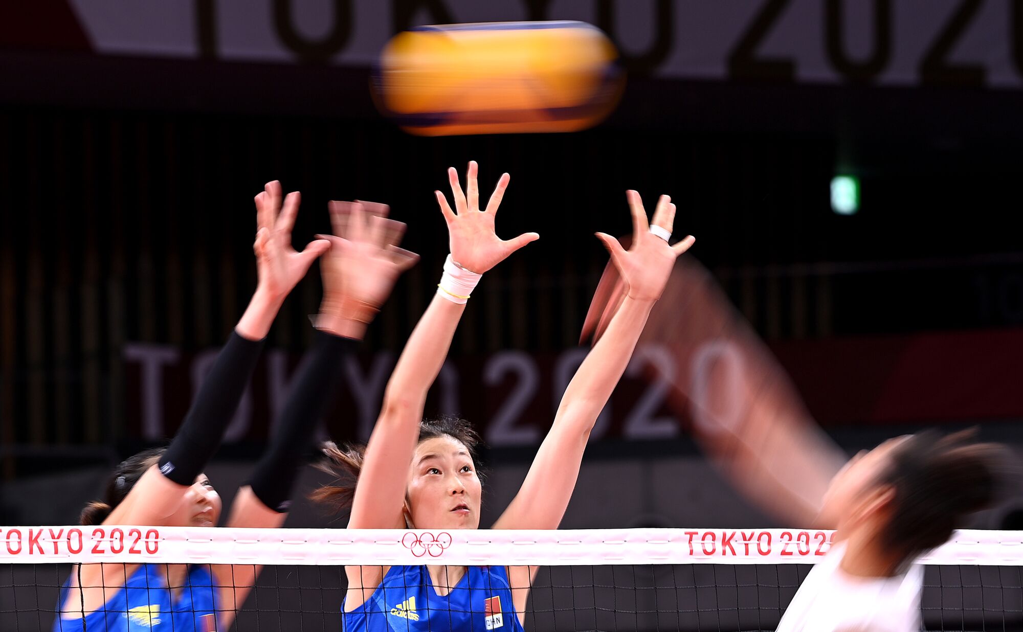 Three women reach up at the net; the volleyball is a blur among them.