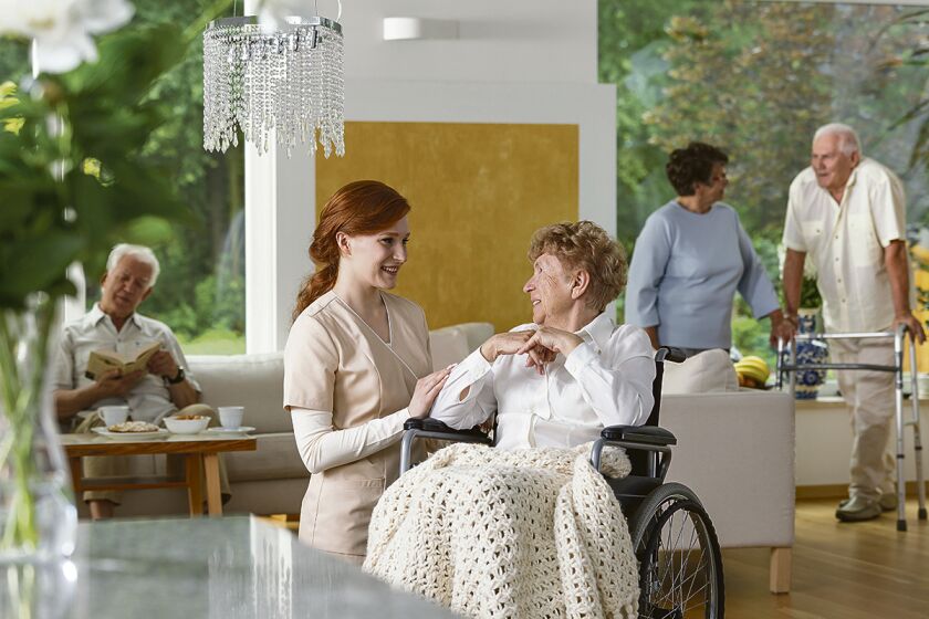 Seniors in a luxury living room of a private retirement home. Tender caregiver by an elderly lady in a wheelchair in the foreground.