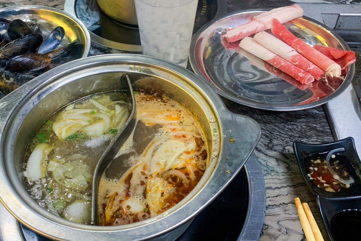 A divided metal pot with two types of soup, next to rolled raw meat on a metal plate