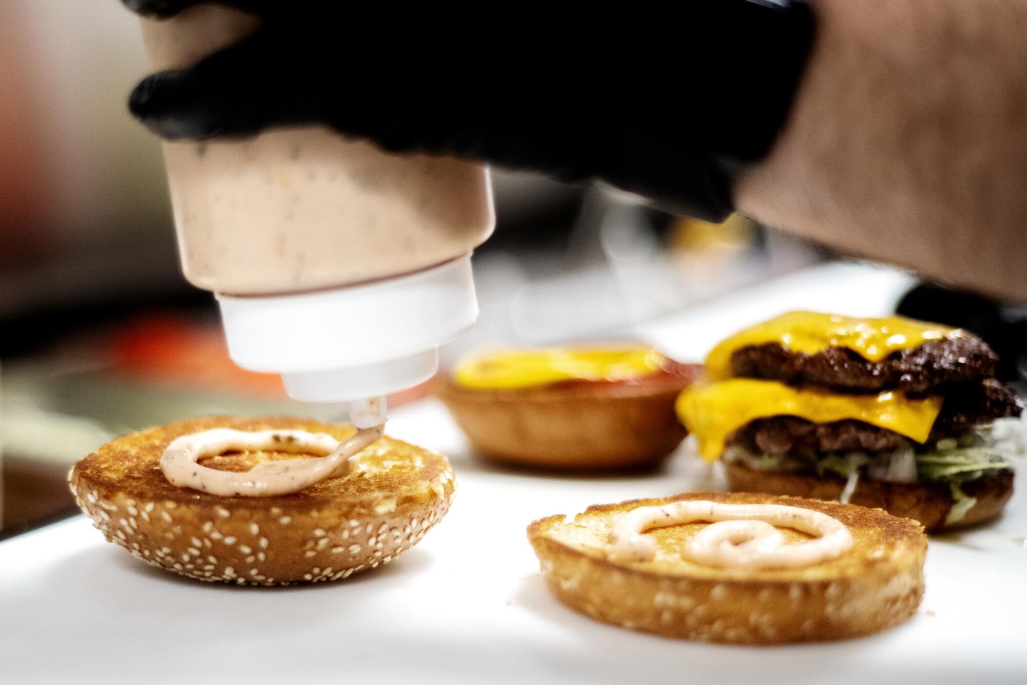 A photo of a hand squeezing a bottle of house-made sauce onto the tops of burger buns.
