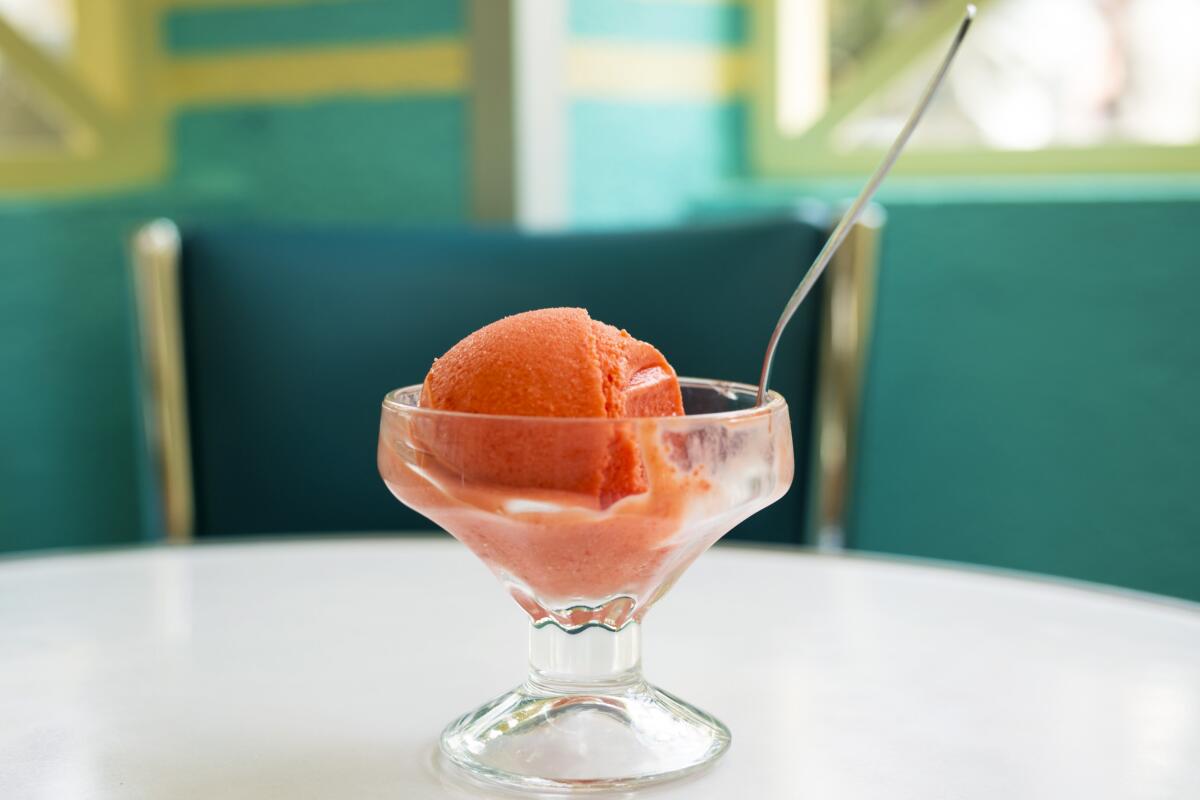 A scoop of pink ice cream sits in a dish with a spoon on a white table.