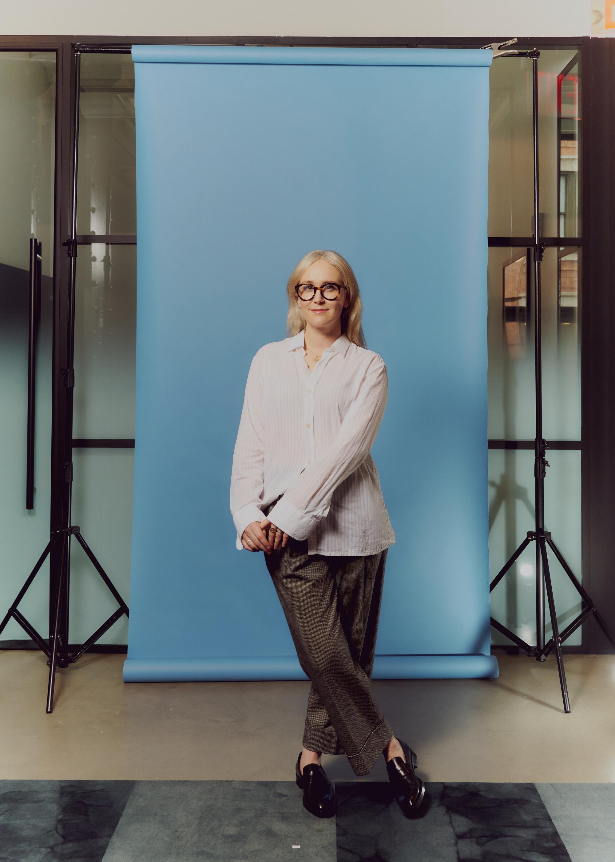 A blond woman wearing glasses, a white button-down shirt and gray trousers, poses in front of a sky blue backdrop. 