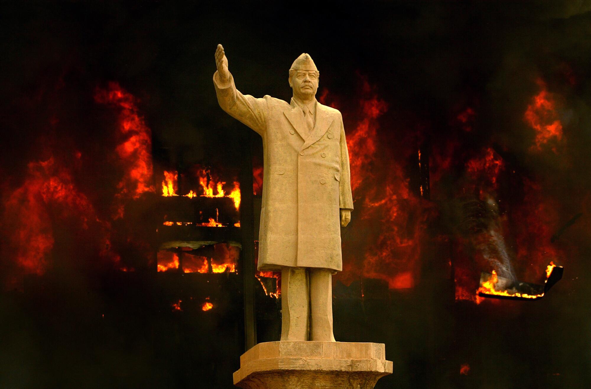 A statue of Saddam Hussein with his arm raised in front of a burning building