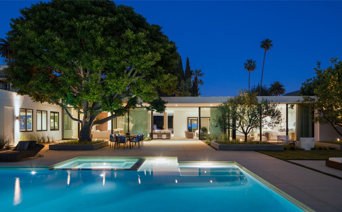 A large pool, outdoor dining area and a low house with glass walls are seen lighted up at night.