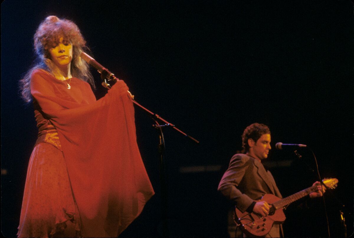 A woman, left, in a flowy red dress holding a microphone and a man playing a guitar onstage