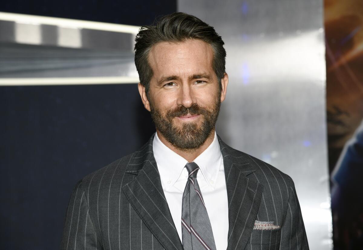 Actor Ryan Reynolds with facial hair in a black striped suit and a silver tie