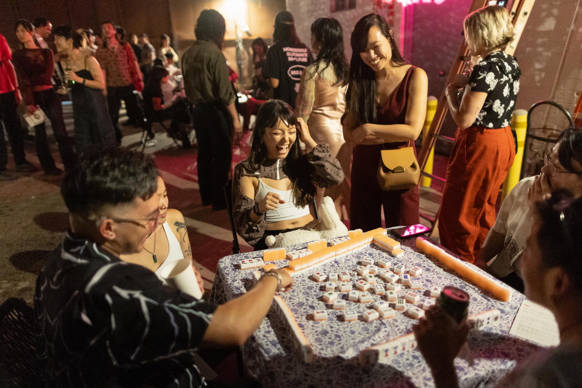 A woman looks on as four others at a table of four play mah-jongg at a party. 
