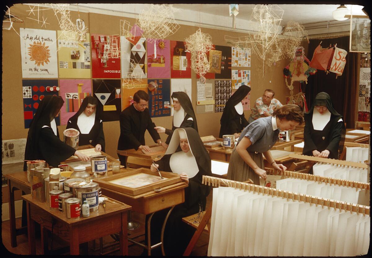 Nuns and a priest work with art materials in a printing studio lined with colorful works