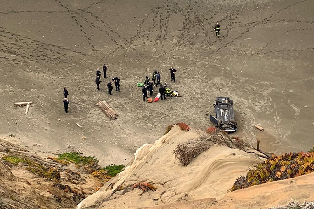 From the top of a cliff, a car is seen upside down in sand, with about a dozen firefighters nearby.