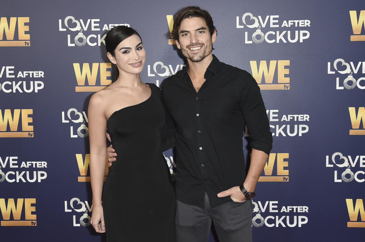 Ashley Iaconetti smiling in a black cocktail dress with Jared Haibon, holding his arm around her and wearing a black shirt.